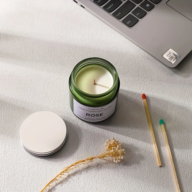 Candle in a green jar labeled 'Rose' on a light surface with a laptop and dried flowers.