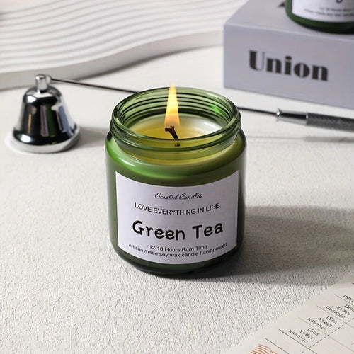 Green tea candle in a glass jar with a lit wick, surrounded by books and stationery items.